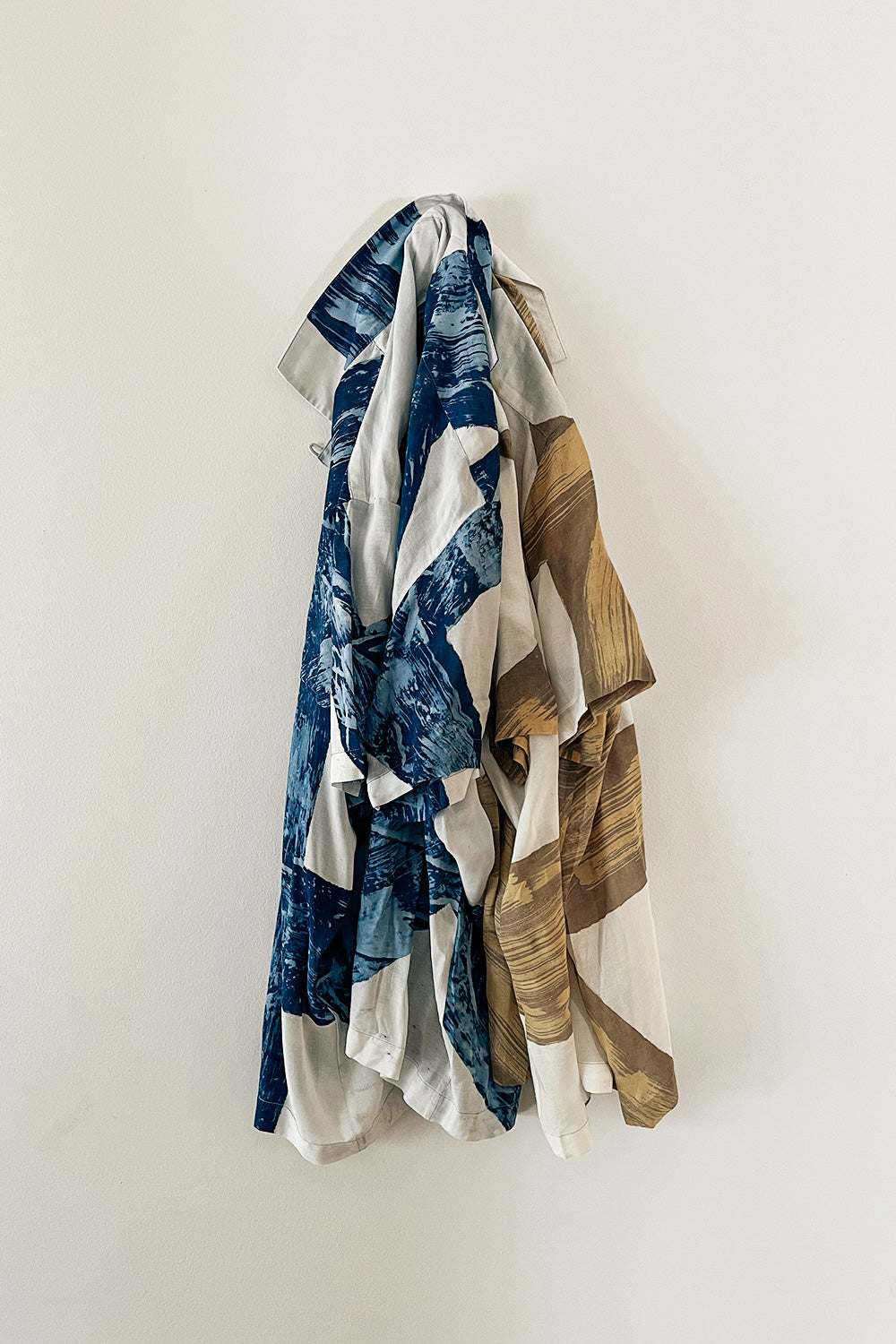 A traditional Alba Batik camp shirt in indigo with a relaxed collar, hanging on a wall. The shirt features a geometric pattern with blue and brown colors.