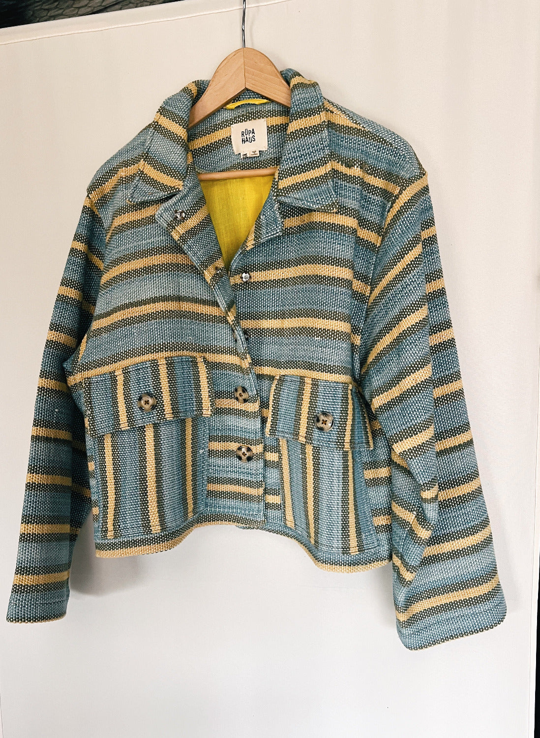 A striped multicolored jacket with asymmetrical deep pocket placement and button detailing, hanging on a hanger against a white background.
