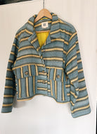 A striped multicolored jacket with asymmetrical deep pocket placement and button detailing, hanging on a hanger against a white background.