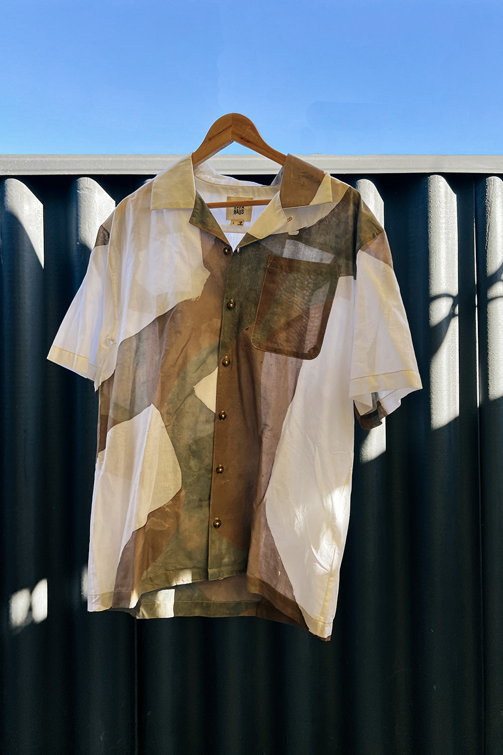 A handpainted batik cotton shirt in beige, brown, and white colors, displayed on a hanger against a blue background, with a camouflage pattern.