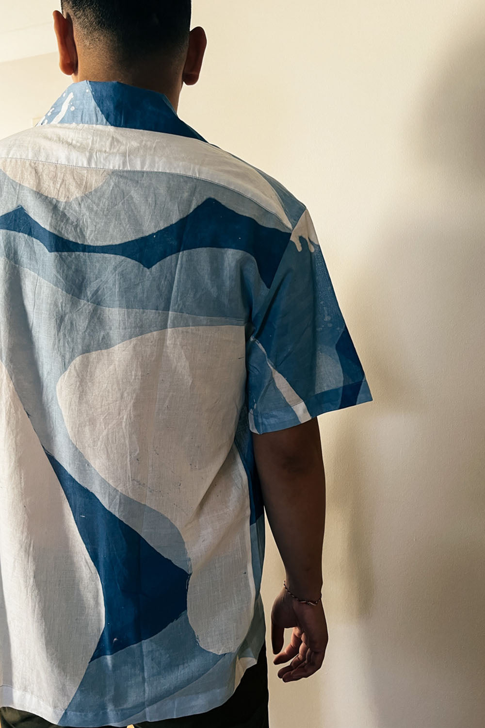 A handpainted batik cotton shirt with a relaxed collar, featuring indigo and natural hues with contrasting color buttons and a chest pocket, worn by a male model.