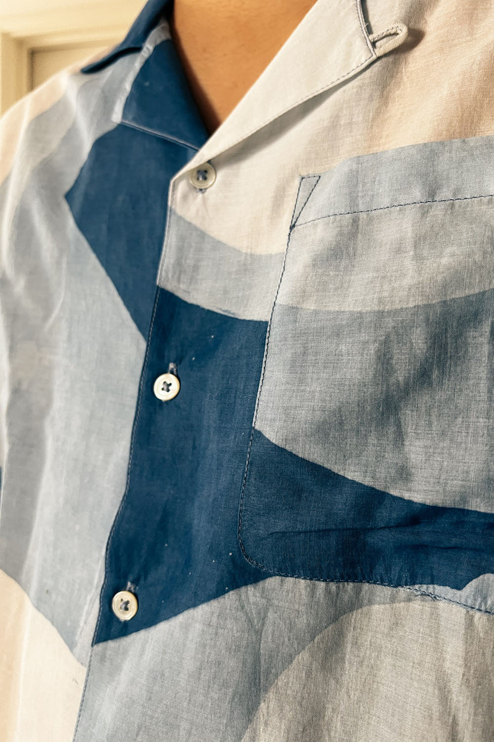 A handpainted batik cotton shirt with a relaxed collar, featuring indigo and natural hues with contrasting color buttons and a chest pocket, worn by a male model.