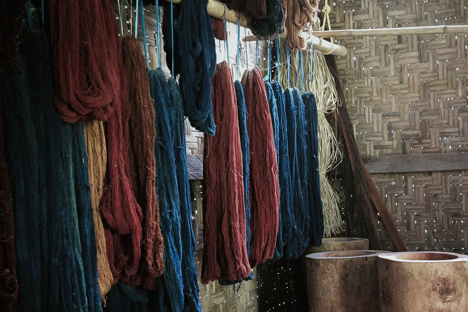 Handwoven cotton yarns dyed with plant-based pigments hanging beside wooden dye vats in an Indonesian workshop, showing natural variation from small-batch, manual dyeing processes.
