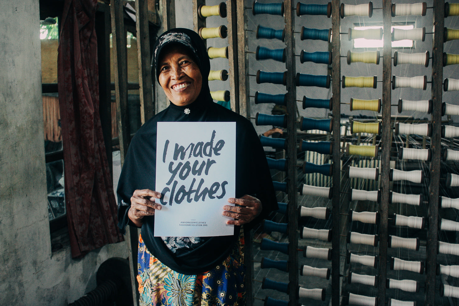 Lorek weaver holding a “Who Made My Clothes?” sign as part of a transparency campaign, photographed outside a workshop