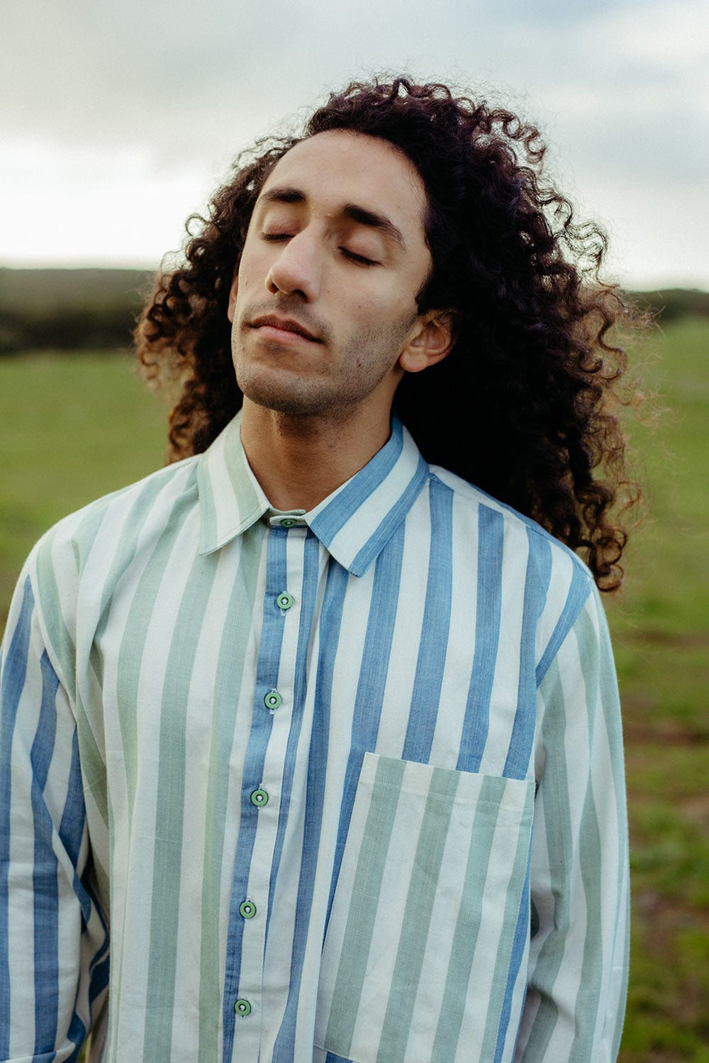 A person with a contemplative expression wearing a striped blue and green shirt with a classic collar and structured cuffs.