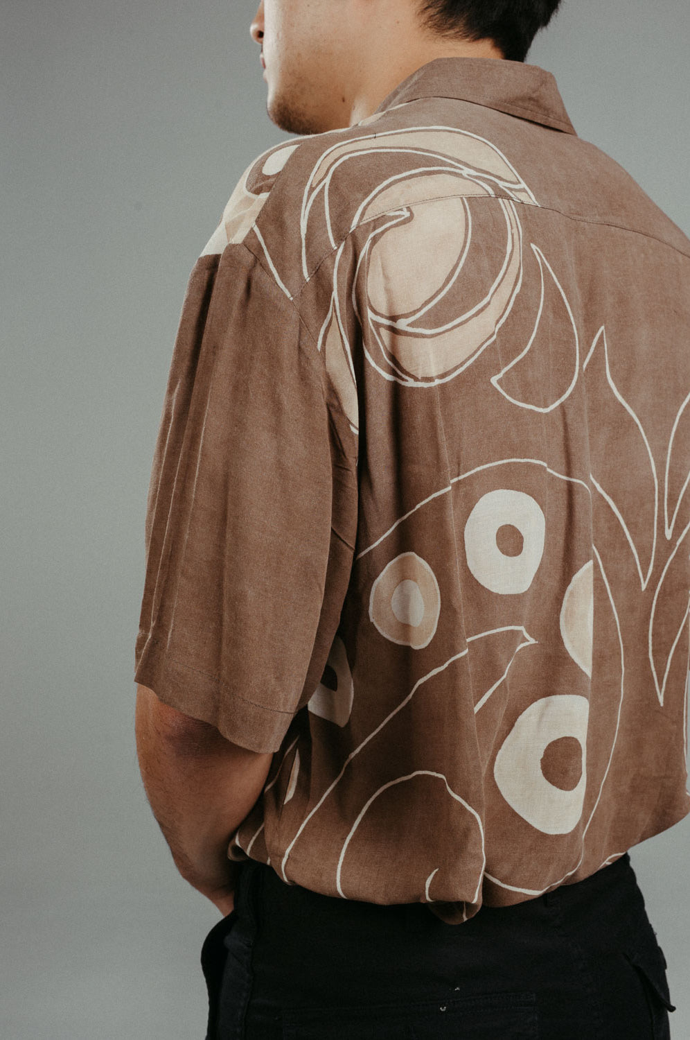 A person wearing a brown handpainted batik shirt with geometric patterns and a classic collar, featuring contrasting recycled seashell marble buttons.