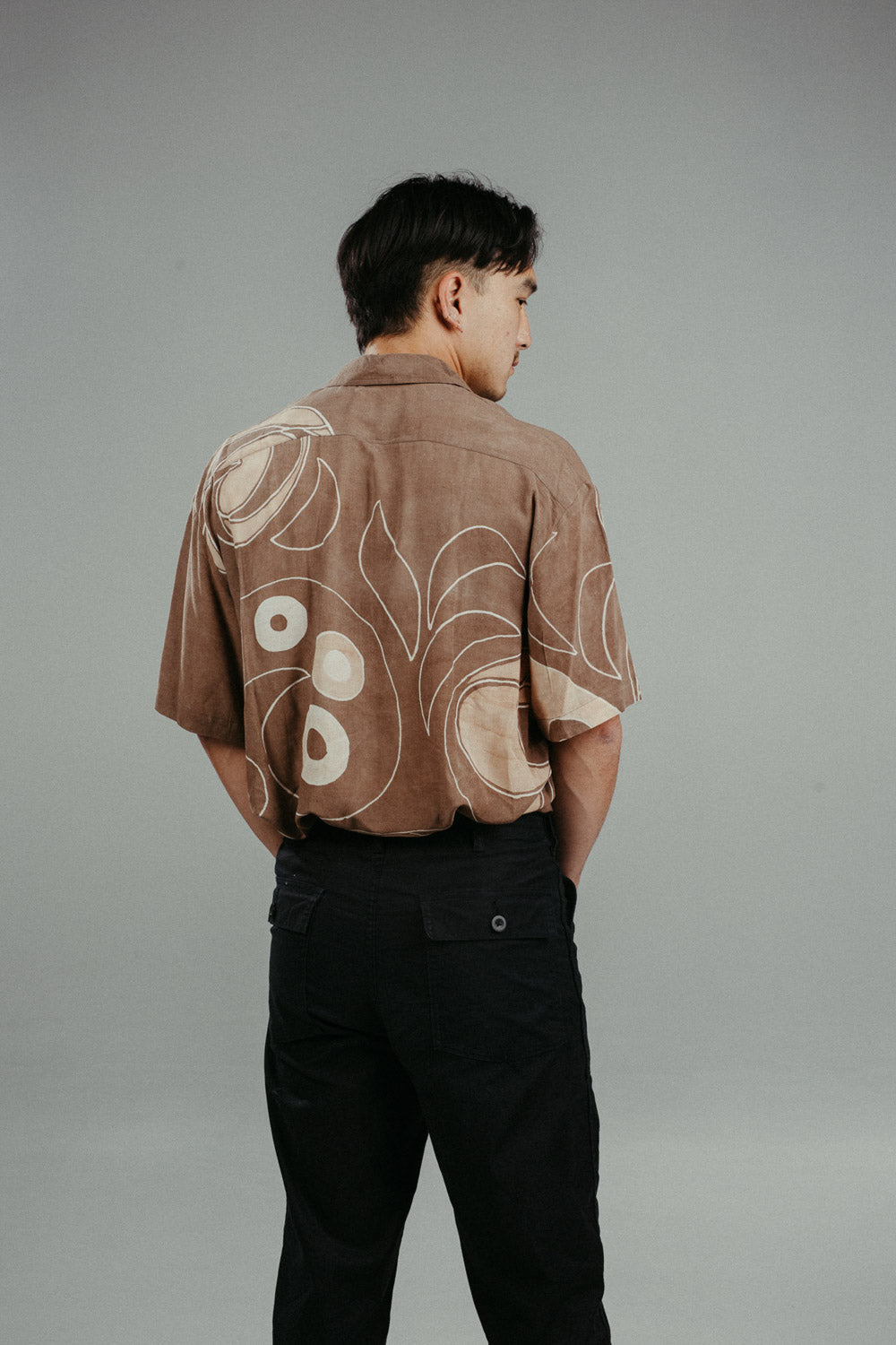 A person wearing a brown handpainted batik shirt with geometric patterns and a classic collar, featuring contrasting recycled seashell marble buttons.
