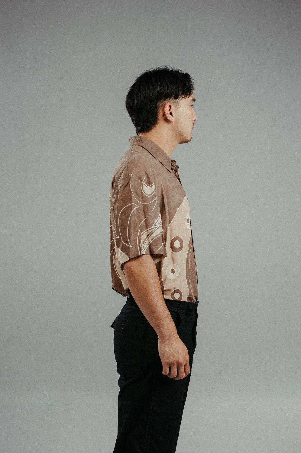 A person wearing a brown handpainted batik shirt with geometric patterns and a classic collar, featuring contrasting recycled seashell marble buttons.