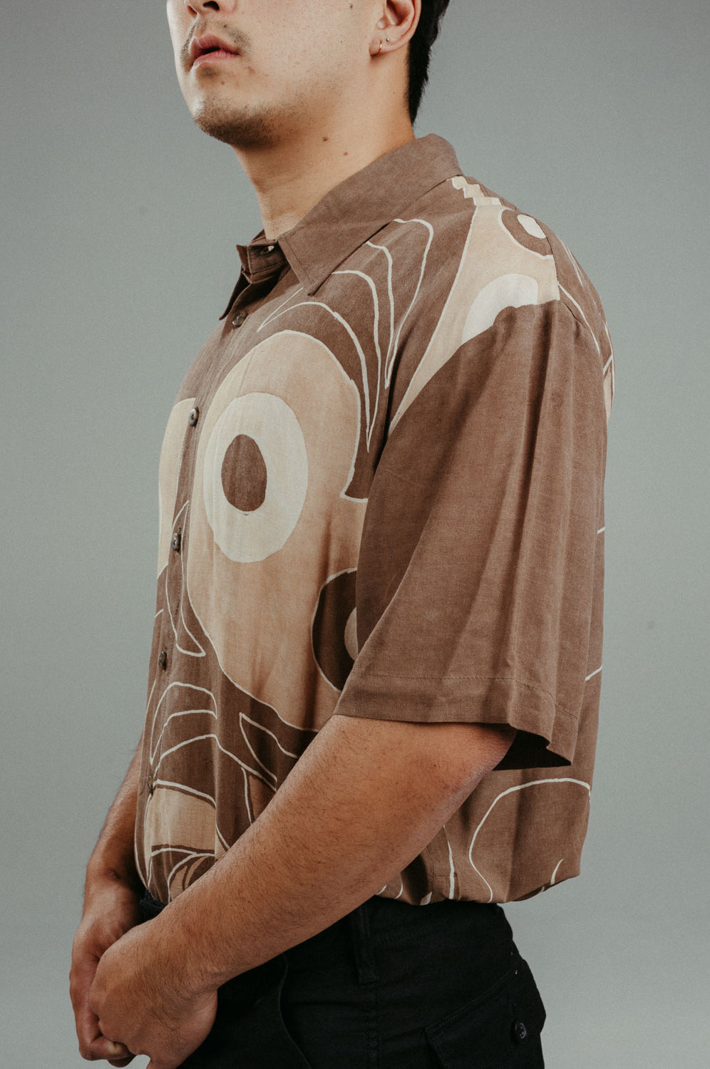 A person wearing a brown handpainted batik shirt with geometric patterns and a classic collar, featuring contrasting recycled seashell marble buttons.