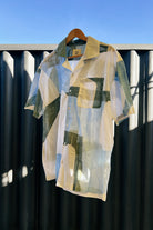 Handpainted Batik Camp Shirt in blue, green and natural hues with relaxed collar and short sleeves, hanging on a wooden hanger against a metal fence background.