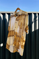 A multicolored batik camp shirt with a relaxed collar and contrasting color buttons, hanging on a hanger against a metal fence background.