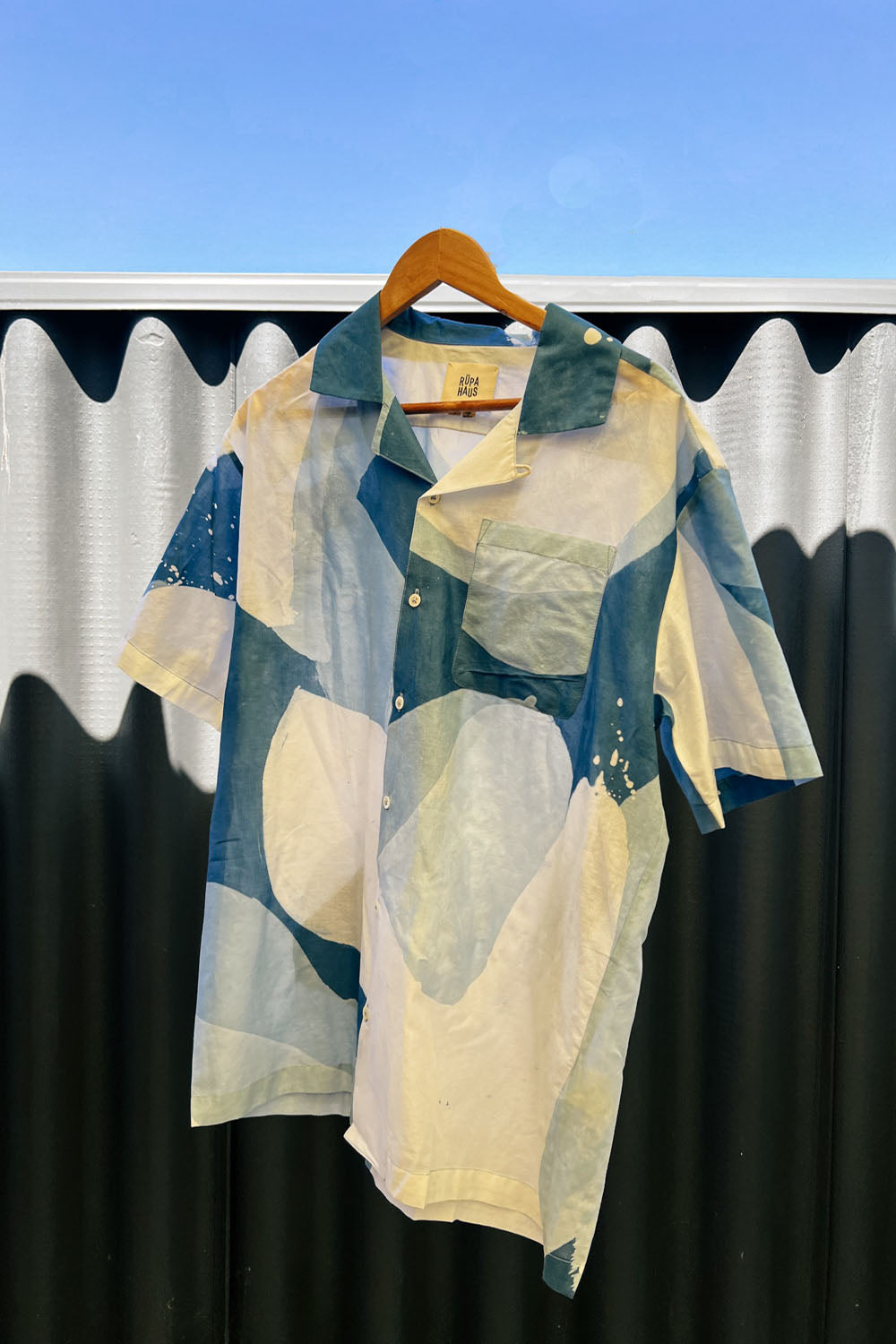 A handpainted batik cotton shirt with a relaxed collar, featuring indigo and natural hues with contrasting color buttons and a chest pocket, displayed on a hanger.