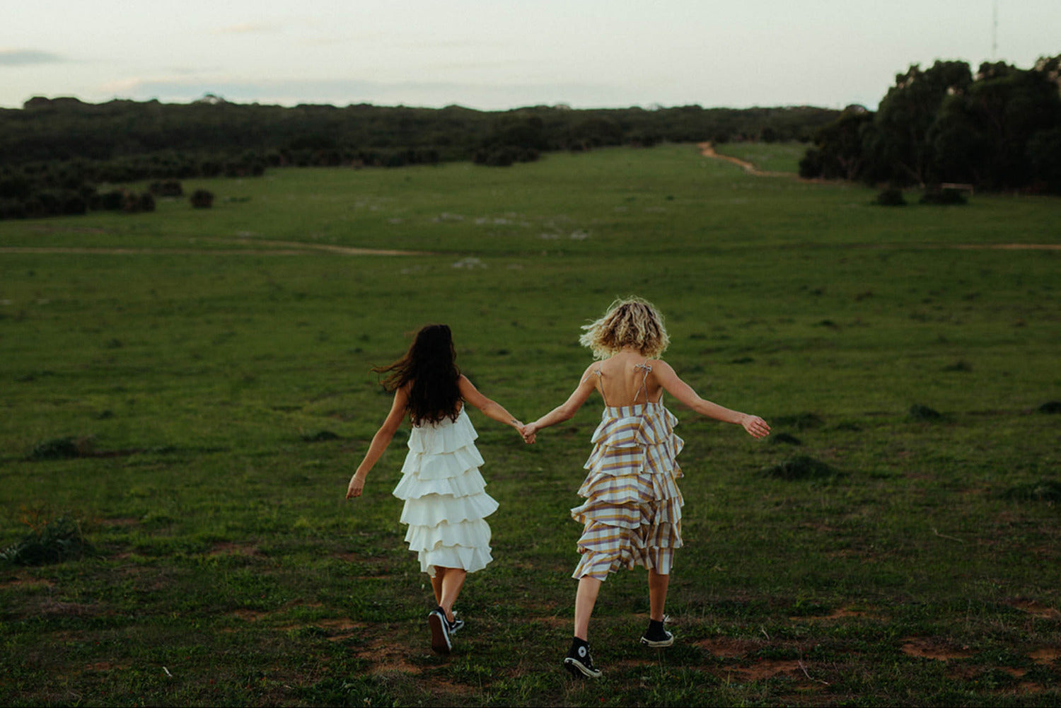 Two women walking hand in hand across an open field wearing layered, seasonless RŪPAHAUS dresses designed for everyday wear and long-term use.