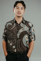 A person wearing a brown handpainted batik shirt with a geometric pattern featuring circles and lines.