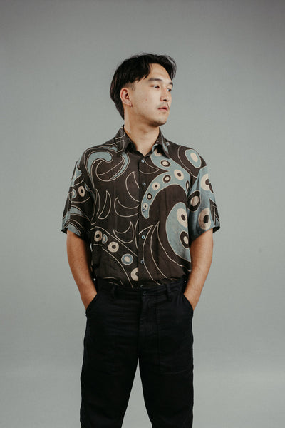 A person wearing a brown handpainted batik shirt with a geometric pattern featuring circles and lines.