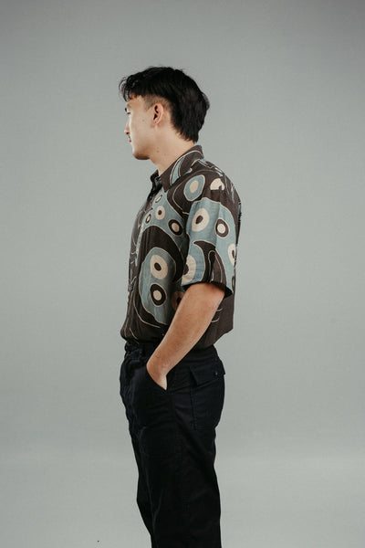 A person wearing a brown handpainted batik shirt with a geometric pattern featuring circles and lines.