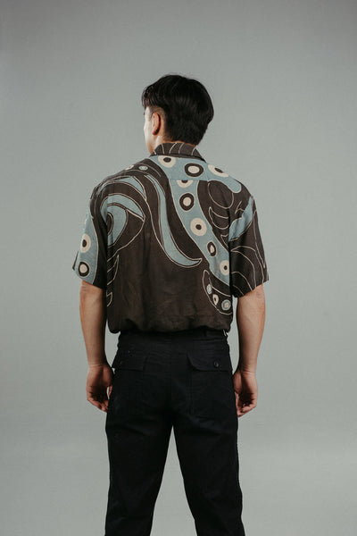 A person wearing a brown handpainted batik shirt with a geometric pattern featuring circles and lines.
