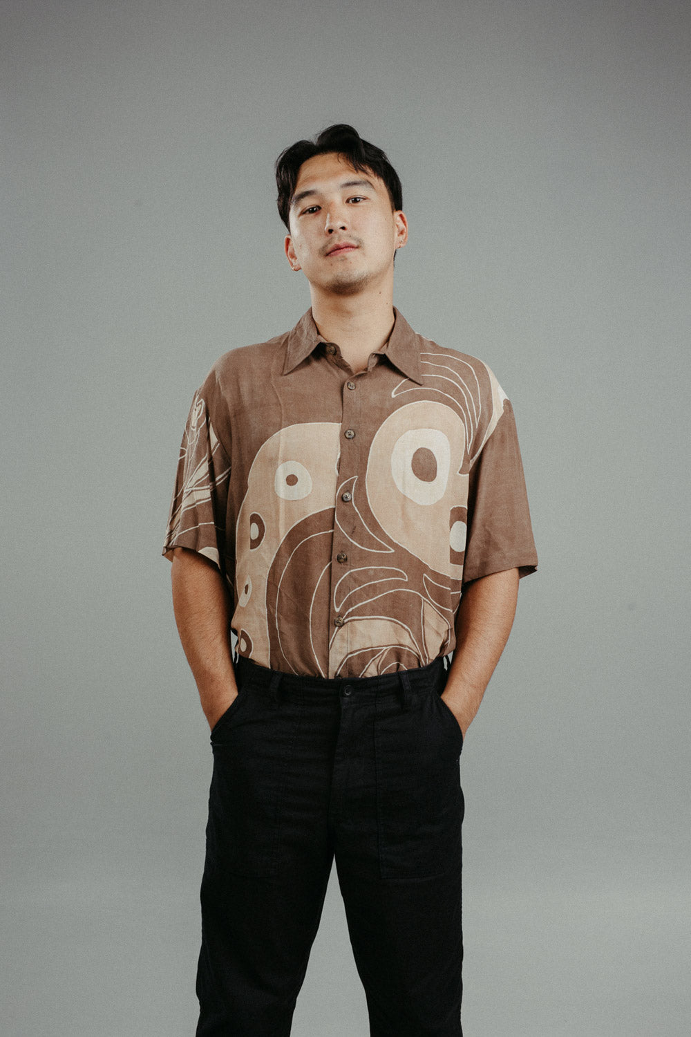 A person wearing a brown handpainted batik shirt with geometric patterns and a classic collar, featuring contrasting recycled seashell marble buttons.